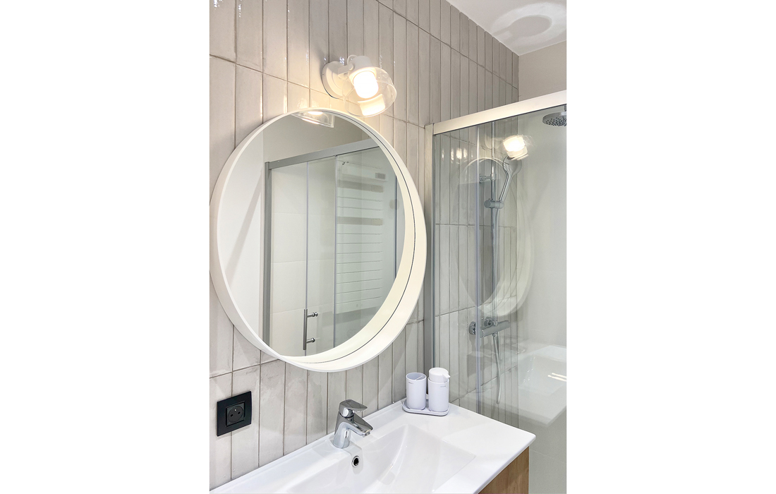 détail meuble vasque d'une salle d'eau avec une faience beige en pose verticale un meuble bois vasque incruste un miroir rond blanc et une applique transparente